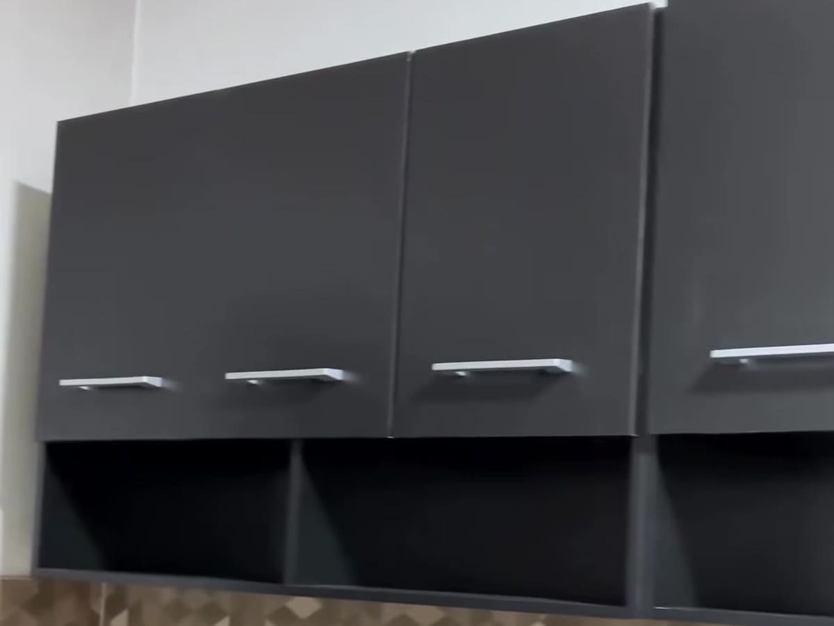 Armários aéreos de cozinha em cinza fosco, quatro portas com puxadores prateados e nichos abertos embaixo, presos na parede acima de um revestimento com desenho de cubos.