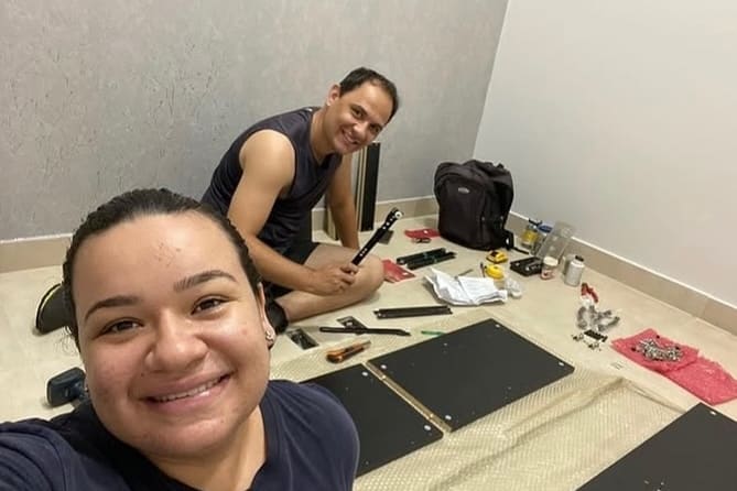 William e Ariane montando um móvel no chão de um quarto: ela sorri para a câmera em primeiro plano, ele está sentado atrás com uma corrediça de gaveta na mão, entre as peças pretas do móvel abertas sobre plástico-bolha, as ferramentas e os parafusos separados.