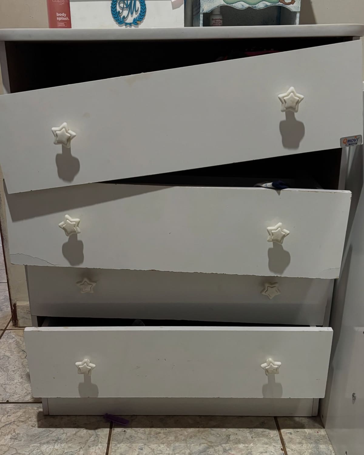 Cômoda infantil branca de puxadores em formato de estrela: a gaveta de cima está atravessada e saindo do corpo do móvel, a do meio tem a frente rachada de ponta a ponta e a de baixo está para fora, desencaixada.