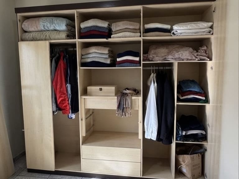 Interior de um guarda-roupa de madeira clara com as portas abertas: prateleiras com roupa de cama e agasalhos dobrados, duas araras com camisas e casacos pendurados e gavetas na parte de baixo.