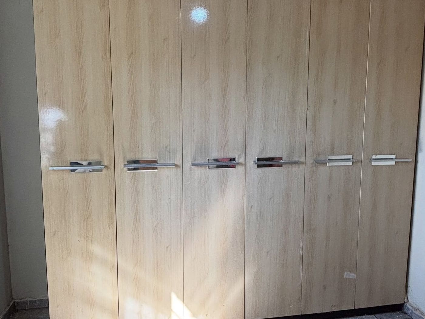 Guarda-roupa de seis portas em madeira clara, com puxadores prateados na altura da cintura, encostado na parede de um quarto vazio.